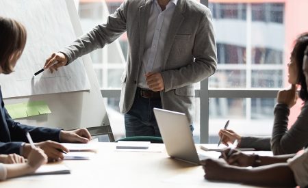 Skilled 30s caucasian leader in eyeglasses coaching interested business people, giving educational workshop presentation in office. Concentrated employees listening to professional coach at workshop.